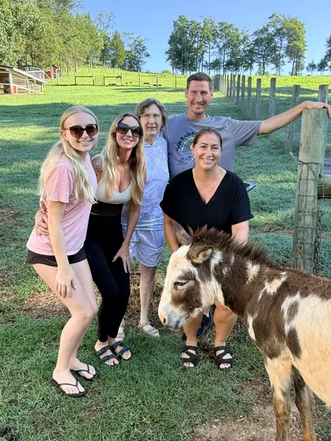 A group of five visitors smiling by the fence line with a brown-and-white mini donkey in front