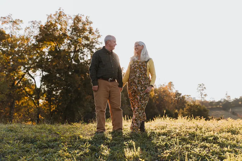 Lane and Jules holding hands in a sunlit field with fall trees behind them
