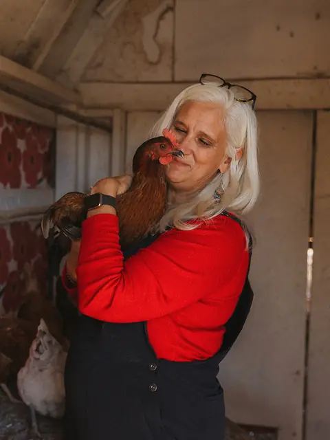 Jules hugging a red hen in the coop, in front of a wall of big red flowers