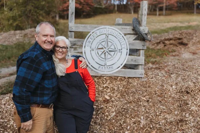 Lane and Jules arm in arm beside the Sweet BombDiggity Farms compass sign on an old wooden gate
