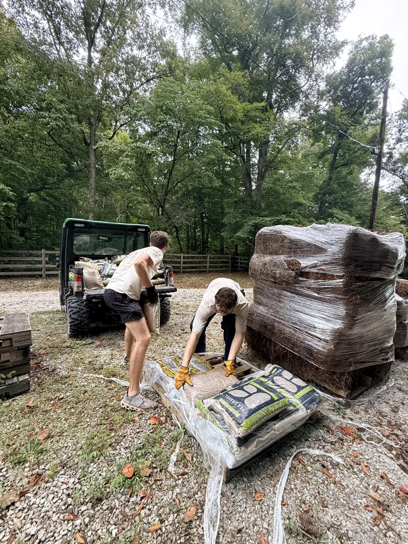 Two volunteers pulling bags of pea gravel off a pallet beside the farm Gator